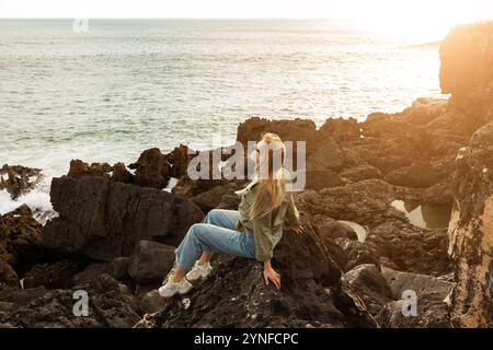 Donna che gode di un tramonto sereno sulla spiaggia rocciosa in una serata tranquilla Foto Stock