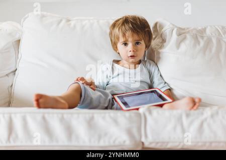 Un bambino si siede comodamente su un divano bianco e si concentra su un tablet. La luce soffusa riempie la stanza, creando un'atmosfera tranquilla come il bambino Foto Stock