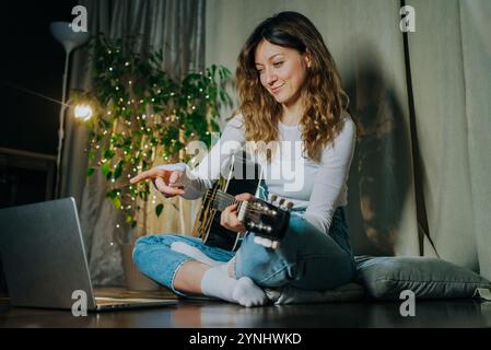 Una giovane musicista sta dando lezioni online di chitarra tramite laptop, seduta sul pavimento a casa, puntando lo schermo del laptop Foto Stock