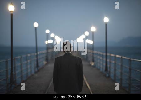 Un uomo anziano con un cappello cammina da solo lungo un molo al crepuscolo. Il soffice bagliore delle lanterne illumina il suo percorso mentre si avvicina alla fine della struttura o Foto Stock