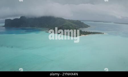 Maupiti, isola a forma di cuore nella Polinesia francese, mostra i suoi vibranti paesaggi verdi e le sue acque incontaminate contro un suggestivo cielo nuvoloso, formando un ipnotico panorama tropicale. Colpo di drone aereo Foto Stock