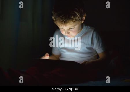 In una stanza buia, un ragazzo siede sul suo letto, assorbito dal bagliore di un tablet digitale. La sua espressione mostra concentrazione e curiosità mentre si impegna Foto Stock