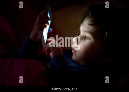 Un bambino osserva con attenzione lo schermo di uno smartphone, illuminato dal suo bagliore in una stanza con luce soffusa. L'atmosfera suggerisce calma e curiosità serali. Foto Stock