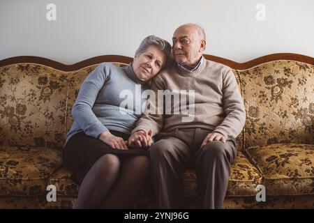 Una coppia di anziani si siede a stretto contatto su un divano vintage floreale, condividendo un momento intimo. Mostrano affetto attraverso gli sguardi morbidi e la mano delicata Foto Stock