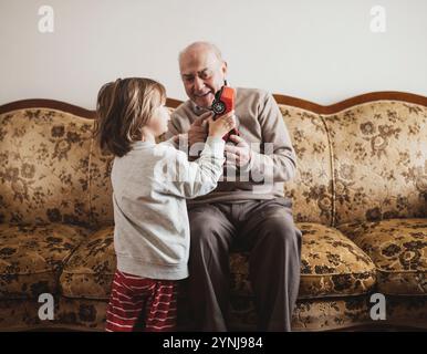 Un bambino gioca con un'auto giocattolo in salotto, sorridendo e condividendo le risate con suo nonno. Entrambi sono comodamente seduti su un divano decorato, Foto Stock