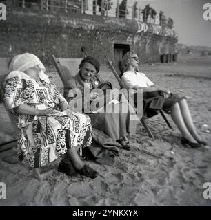 Anni '1950, storico, tre donne sedute su sdraio su una spiaggia sabbiosa, Inghilterra, Regno Unito. Due sono anziani, uno ha un cappotto, mentre l'altro indossa un lungo abito a motivi geometrici ha una sciarpa sopra la testa e ha una doze. Una ragazza più giovane in una sdraio accanto a loro, in una gonna e in un top, sta dormendo. Foto Stock