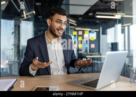 Uomo d'affari in un ufficio moderno che partecipa a riunioni virtuali tramite laptop. Espressione sicura, gesti aperti mettono in risalto la comunicazione e la professionalità. SMA Foto Stock