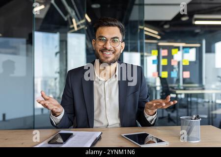 Un uomo d'affari fiducioso in un ufficio moderno sorridente calorosamente e impegnato in una conversazione. Tecnologia come telefono e tablet presente sulla scrivania, che riflette i professi Foto Stock