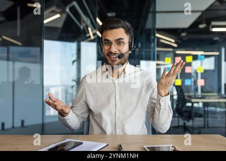 Uomo sicuro di sé che indossa una cuffia e che partecipa a una videochiamata in un ufficio moderno. Mostra entusiasmo ed energia. Circondato da strumenti digitali l Foto Stock