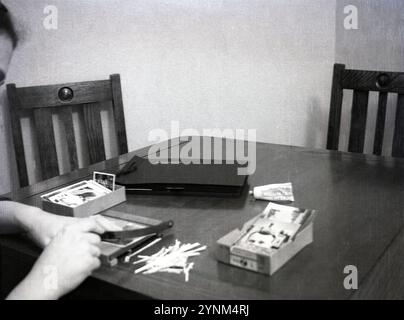 Anni '1950, fotografia storica, di casa, seduto a un tavolo di legno, un uomo che usa un taglierino di carta o una mini ghigliottina, per tagliare i lati delle foto che metterà in un album fotografico, Inghilterra, Regno Unito. Nei giorni precedenti la fotografia digitale, le fotocamere utilizzavano pellicole per catturare immagini e visualizzare le immagini le stampe dovevano essere sviluppate in speciali laboratori cinematografici dai negativi. Foto Stock
