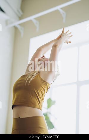 La donna si allunga graziosamente in un luminoso studio, incarnando equilibrio e pace durante la pratica yoga. I suoi capelli biondi cadono dolcemente mentre emana serenità e calma, mostrando flessibilità e forza Foto Stock