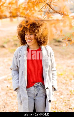 Una donna allegra si erge tra foglie d'autunno dorate, indossando un comodo cappotto grigio e un maglione rosso. Il suo sorriso luminoso riflette il calore e la bellezza del Foto Stock