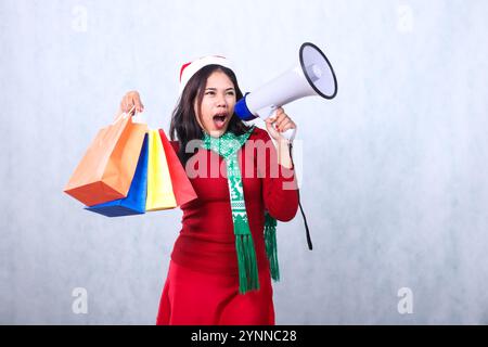 affascinante donna adulta che indossa un maglione rosso natalizio, un cappello e una sciarpa di babbo natale, che urla arrabbiata mano schietta che tiene il megafono e solleva il colorfu Foto Stock