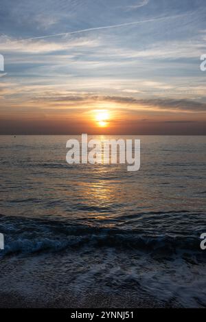 Scatto verticale di un tramonto in mare con la silhouette di un faro all'orizzonte, di colore argento e arancione. Foto Stock
