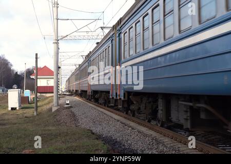 Un treno blu passa davanti a una pittoresca stazione immersa in un paesaggio rurale. Il sole proietta un caldo bagliore sull'ambiente circostante, mettendo in risalto l'atmosfera tranquilla Foto Stock