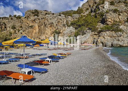 Spiaggia di ghiaia con lettini e ombrelloni colorati, circondata da scogliere rocciose, Ahata Beach, Ahata, East Coast, Karpathos, Dodecaneso, isola greca Foto Stock