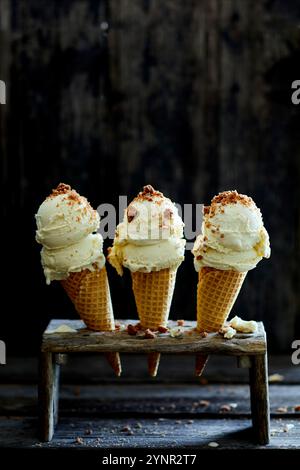 tre coni di gelato ripieni di gelato alla vaniglia e briciole di biscotti, in un banco di gelato di legno, su uno sfondo di legno scuro Foto Stock