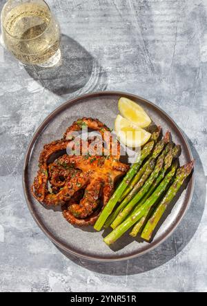 Delizioso polpo alla griglia servito con vivaci lance di asparagi e spicchi di limone fresco su un piatto grigio. Il sole schiarisce immitazione, duro schiarito. Foto Stock