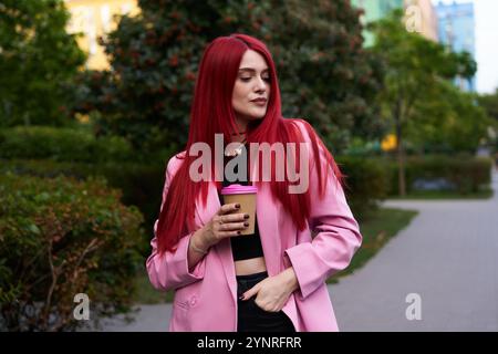 Una donna dai capelli rossi accesi si gusta il caffè in un vivace parco urbano Foto Stock