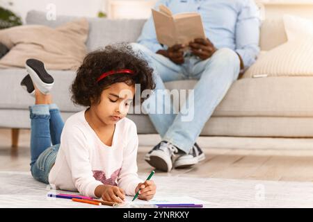 Momenti di tranquillità a casa. Bella ragazza che disegnava sul pavimento e suo nonno che legge interessante libro in soggiorno Foto Stock