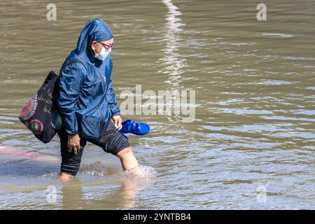 SAMUT PRAKAN, THAILANDIA, 19 novembre 2024, una donna anziana cammina con attenzione attraverso una strada allagata Foto Stock