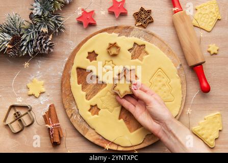 Donna che prepara i biscotti di natale utilizzando taglierine per biscotti di forma diversa su un asse di legno, circondato da decorazioni festose Foto Stock