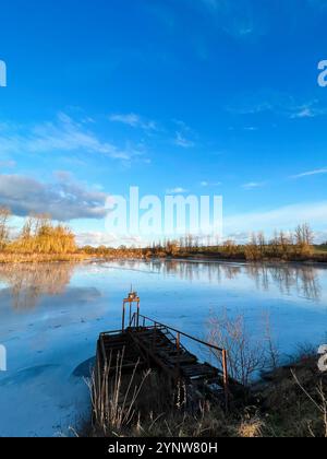 Un lago ghiacciato riflette il sereno cielo invernale con un molo in legno invecchiato solitario che si protende. Gli alberi senza foglie circostanti sono immersi nel caldo bagliore di Foto Stock