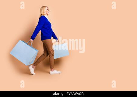 Elegante donna matura con capelli biondi che trasporta borse della spesa su sfondo beige con eleganti abiti e occhiali Foto Stock