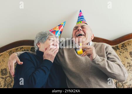 Una coppia di anziani si siede insieme su un divano, indossando cappelli colorati da festa. Condividono un momento di risate, mostrando la loro felicità e connessione du Foto Stock