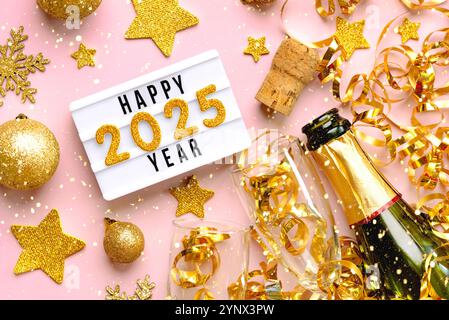 Felice anno nuovo 2025. Vista dall'alto della scatola luminosa con la parola "anno felice 2025", palline di natale dorate, bicchieri di cristallo e bottiglia di champagne sul backgroun rosa Foto Stock