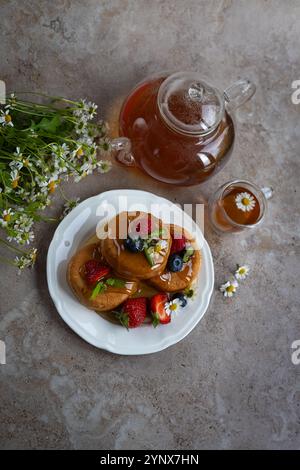 Deliziosa pila di soffici pancake conditi con miele, conditi con frutti di bosco freschi e foglie di menta, serviti con tè di camomilla in una teiera di vetro Foto Stock