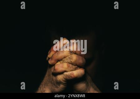 Primo piano di mani maschili disperatamente unite in un gesto di preghiera o di contemplazione profonda con sfondo scuro. Foto Stock