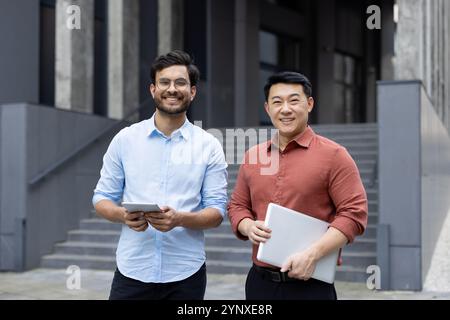 Professionisti aziendali sicuri che si trovano all'aperto con dispositivi digitali, sorridenti in un ambiente urbano moderno. Uomini che mostrano il lavoro di squadra e la collaborazione nell'ambiente aziendale. Foto Stock