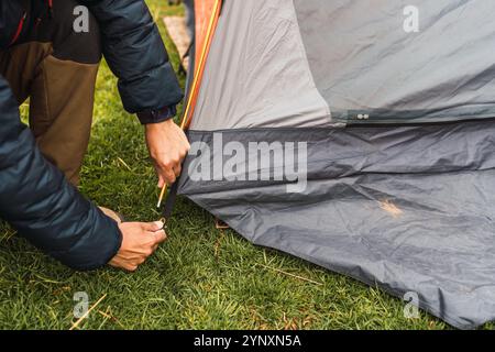 Le mani dell'uomo sistemano una tenda arancione sull'erba verde illuminata dalla luce naturale nella catena montuosa delle Ande Foto Stock