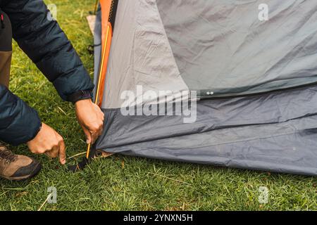 Le mani dell'uomo sistemano una tenda arancione sull'erba verde illuminata dalla luce naturale nella catena montuosa delle Ande Foto Stock