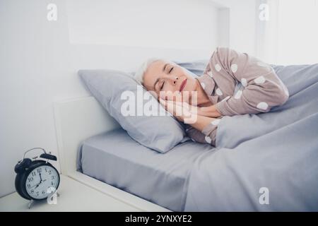 Donna anziana che dorme tranquillamente nel letto indossando pigiama a pois, abbracciando serenità e relax a casa con luce naturale Foto Stock