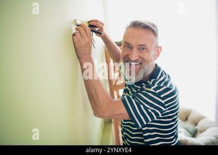 Uomo anziano che trascorre un weekend rilassante a casa dipingendo, sorridendo in una t-shirt casual all'interno, catturando momenti di gioia alla luce naturale del giorno Foto Stock