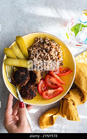 Vista dall'alto di una donna che mangia grano saraceno, cotolette di carne, pomodori e cetriolini sottaceto con un bicchiere di acqua ghiacciata e menta fresca Foto Stock