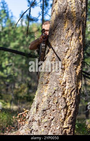 Hunter si nasconde dietro un grande albero e punta un fucile mentre guarda attraverso il mirino. Pronti a sparare. Caccia nella foresta. Vista frontale. Foto Stock