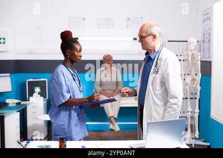Il medico maschio anziano e l'assistente medico femminile sono approfonditi nella discussione sulla salute del paziente. Infermiera nera con appunti, in piedi in stanza clinica con medico in pensione e scrivere istruzioni mediche. Foto Stock