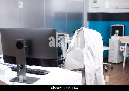 In ufficio medico, camice bianco da laboratorio posto su sedia vuota con scrivania che tiene il pc desktop e la tastiera, evidenziando una postazione di lavoro preparata per il medico. Foto Stock