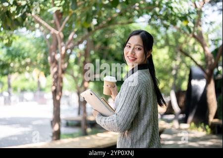 Una giovane studentessa asiatica, affascinante e felice, in abiti caldi, porta con sé la sua bevanda calda e gli elementi essenziali della scuola, cammina lungo il sentiero del campus Foto Stock