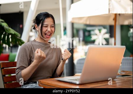 Una allegra donna asiatica seduta davanti al suo laptop all'aperto, guardando lo schermo con un'espressione felice, alza il pugno di trionfo, celebrando lui Foto Stock