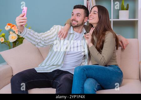 Coppia che scatta selfie sul divano nell'accogliente soggiorno con smartphone e sorride. Foto Stock