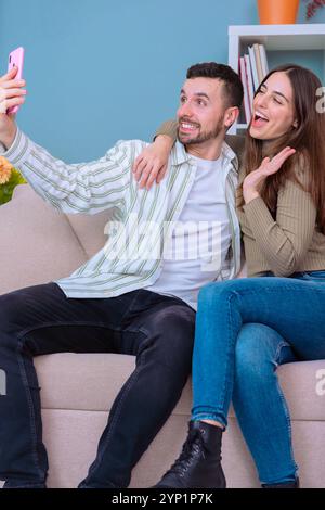 Coppia che scatta selfie sul divano nell'accogliente soggiorno con smartphone e sorride. Foto Stock