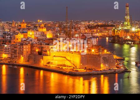 Vista panoramica notturna della storica città fortificata di Birgu (Vittariosa) con piattaforma petrolifera e altri impianti industriali attraverso il Grand Harbor da va Foto Stock