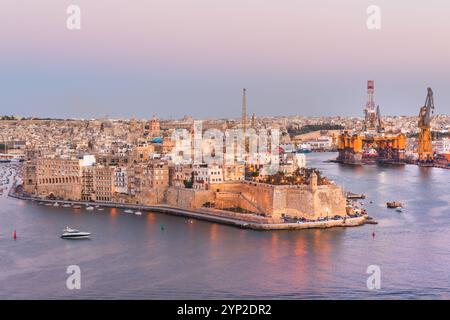 Vista panoramica al tramonto della storica città fortificata di Birgu (Vittariosa) con piattaforma petrolifera e altri impianti industriali attraverso il Grand Harbor dalla Valle Foto Stock