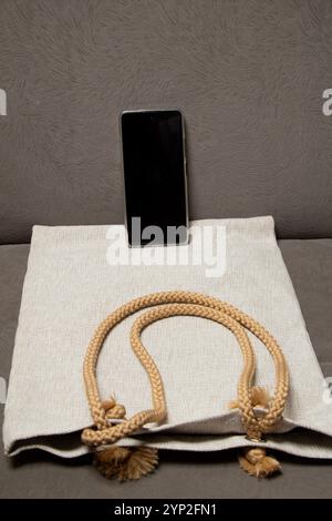 Ritratto della borsa tote Jute Wick con schermo del telefono bianco e nero su divano grigio, eco-friendly beige Label Mockup Foto Stock