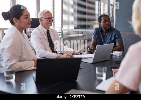 Medico afroamericano maschile in scrub blu impegnato in discussione di gruppo con i colleghi su anamnesi di pazienti malattia, mentre seduto a meeti Foto Stock