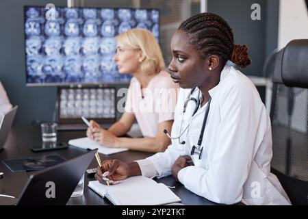 Vista laterale della radiologa afroamericana che ascolta le opinioni dei colleghi durante una discussione di gruppo mentre scrive appunti su un notebook seduto in meeti Foto Stock